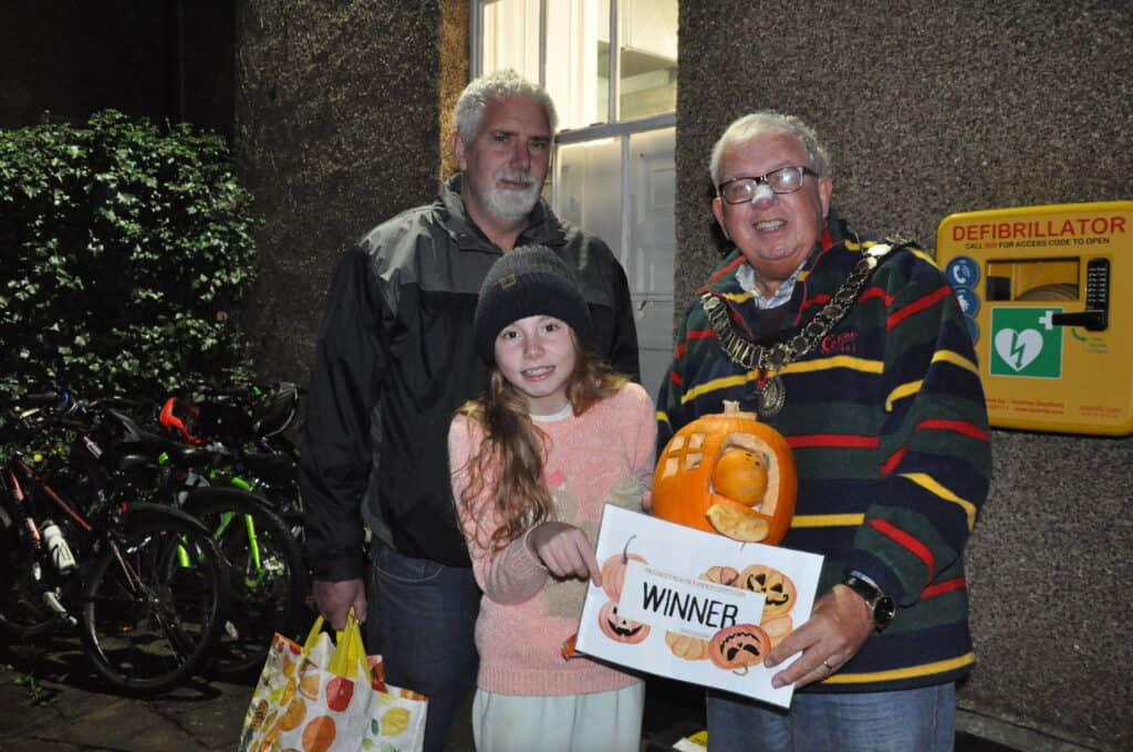 The Mayor of Sedgefield Peter Hinde standing with children's pumpkin competition winner Elsie and a family member, displaying the winning pumpkin and certificate. The pumpkin is shaped like a house, with a window and doorway cut out, with some pumpkin slice steps and a happy little pumpkin wedged in the doorway.