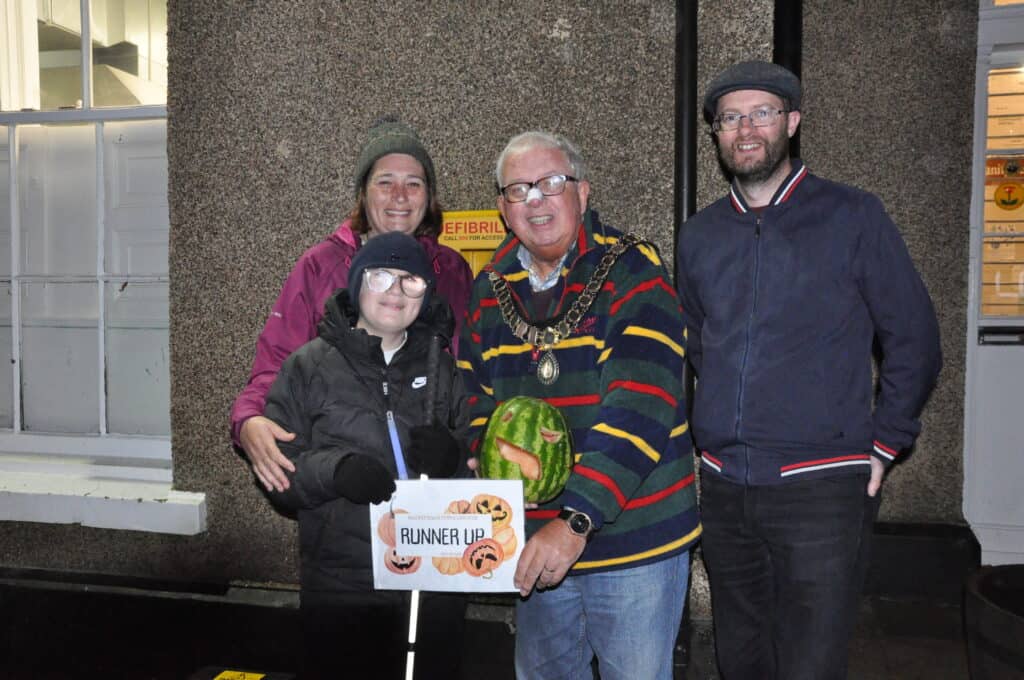 The Mayor of Sedgefield Peter Hinde standing with children's pumpkin competition runner up Olly with his parents, displaying his watermelon with a carved face and the runner up certificate. 